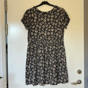 Blommig kortklänning med dragkedja - Supersöt klänning med vita och gula blommor och små rutiga detaljer. Klänningen har korta ärmar, rund halsringning och en synlig dragkedja i ryggen. Perfekt för dig som gillar en lekfull och avslappnad stil.