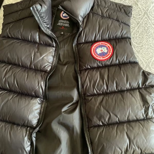 Svart dunväst från Canada Goose - Säljer en svart dunväst från Canada Goose med klassisk logga på bröstet. Västen har quiltad design, hög krage och dragkedja framtill. Perfekt för lager-på-lager och håller dig varm under kyliga dagar.