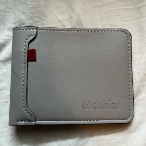 Grå plånbok från dibaoleiou i skinn - Stilren grå plånbok i skinn från dibaoleiou med minimalistisk design. Plånboken har flera kortfack och ett större fack för sedlar. Snygg detalj med en liten röd flik på framsidan. Perfekt för dig som gillar enkel och clean stil.