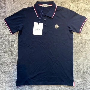 Marinblå moncler piké  - Riktigt schysst marinblå monclear piké med vita och röda ränder i nytt skick. Strl M fast passar även S. En elegant piké som man kan bygga stilfulla outfits med utan problem, hör av er vid frågor!                                     ⚜️Gratis armband vid köp av detta plagg. Välj mellan ett Anchor eller cuff armband, annars medföljer ett slumpmässigt⚜️