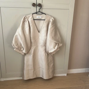 Champagnefärgad klänning från Joelle  - Superfin kort klänning i champagnefärgat satin från Joelle. Klänningen har puffiga trekvartsärmar och djup v-ringning framtill. Snygg minimalistisk design med lyxig känsla och lätt glansigt material. Perfekt för dig som vill sticka ut med stil.