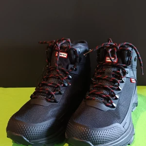 Hunter svarta kängor med snörning - Säljer ett par svarta Hunter kängor med grov sula och röd logga undertill. Skorna har röd- och svartmönstrade snören, mesh- och syntetmaterial samt robust design för extra grepp. Perfekta för dig som gillar outdoor och vill ha en cool look.