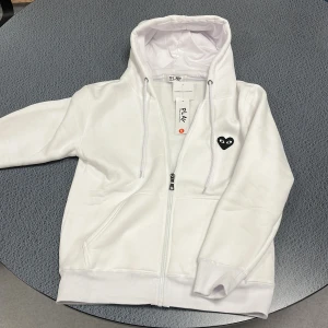Vit hoodie Comme des Garçons Play S - Säljer en clean vit hoodie från Comme des Garçons Play i storlek S. Tröjan har dragkedja, huva med snörning och det ikoniska svarta hjärta-ögon-loggan både fram och bak. Perfekt för dig som gillar stilrena streetwear-plagg.