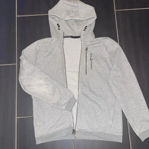 Ljusgrå hoodiejacka från Sail Racing - Ljusgrå hoodiejacka från Sail Racing med dragkedja framtill och bröstficka med logga. Jackan har huva med snörning och ribbade muddar. Perfekt för chill dagar och enkel att matcha med jeans eller joggers.