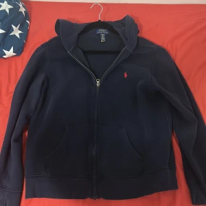 Marinblå hoodie från Polo Ralph Lauren - Snygg marinblå hoodie från Polo Ralph Lauren med röd broderad logga på bröstet. Tröjan har huva, dragkedja framtill och två stora fickor. Tillverkad i mjuk bomullsmix som är perfekt för chill dagar. Klassisk och stilren design som funkar året runt. Storlek XL barnstorlek 18-20 år 