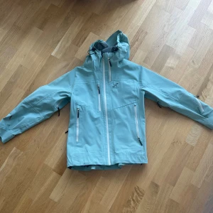 Ljusgrön vindjacka från RevolutionRace - Säljer en ljusgrön vindjacka från RevolutionRace i färgen Iceberg green med huva och flera dragkedjeförsedda fickor. Jackan har justerbar huva, ventilationsdragkedjor under armarna och är tillverkad i slitstark polyester. Perfekt för utomhusaktiviteter och äventyr. Färgen går inte att köpa längre. Väldigt bra skick förutom att jag tejpat om en söm - se bild