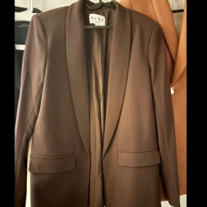 Brun kavaj från NA-KD - Snygg brun kavaj från NA-KD med klassisk krage och två fickor framtill. Stilren och enkel design som passar perfekt till en cool och modern look. Materialet är mjukt och har en lätt glansig finish. Perfekt att styla med jeans eller kostymbyxor.