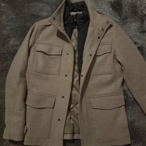 Beige rock från Jack & Jones XS - Snygg beige rock från Jack & Jones Premium i storlek XS. Jackan har fyra stora fickor framtill, hög krage och stängs med både dragkedja och knappar. Insidan är quiltad och ger extra värme, perfekt för kyliga dagar. Stilren design som funkar till många outfits.