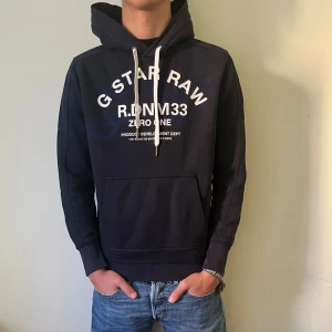 Mörkblå hoodie från G-Star RAW - Snygg mörkblå hoodie från G-Star RAW med vit tryckt logga och text på bröstet. Tröjan passar S-XS. Tröjan har huva med vita snören, stor magficka och ribbade muddar. Perfekt för dig som gillar streetwear och vill ha en chill look.