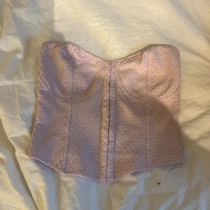 Ljuslila korsett med strukturmönster - Säljer en ljuslila korsett med snygg struktur i tyget och markerade kupor. Toppen är axelbandslös och har en croppad passform som framhäver midjan. Perfekt för dig som vill ha en trendig och feminin look.