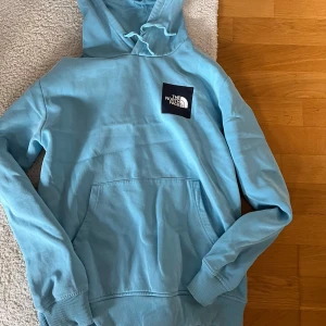Ljusblå hoodie från The North Face - Snygg ljusblå hoodie från The North Face med stor logga på bröstet och coolt grafiskt tryck på ryggen. Tröjan har huva med snörning, magficka och ribbade muddar. Materialet är mjuk bomull och polyester, perfekt för chill dagar. Jag köpä från JD sports 80€