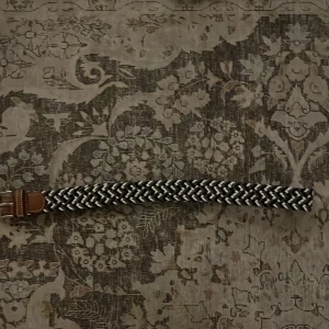 Svart och vit flätad bälte - Säljer ett snyggt flätat bälte i svart och vitt med brunt skinn vid spännet. Bältet har ett randigt mönster och metallspänne. Perfekt för att ge outfiten en cool touch och passar till jeans eller chinos. Flätad design gör det enkelt att justera längden. Den var för lång för mig därför säljer jag den.