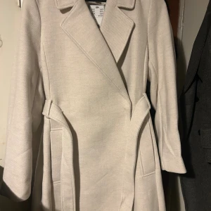 Beige kappa från H&M med bälte - Snygg beige kappa från H&M med klassisk krage och bälte i midjan. Långärmad och med stilren design, perfekt för dig som gillar cleana och neutrala looks. Materialet är mjukt och känns lyxigt, och kappan har praktiska fickor på sidorna.