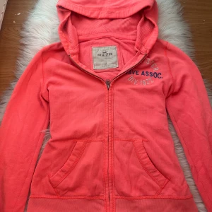 Korallrosa hoodie från Hollister XS - Snygg korallrosa hoodie från Hollister med dragkedja, huva och fickor framtill. Tryck med Hollister Wave Assoc. på bröstet och ärmen. Perfekt för chill dagar och enkel att matcha med jeans eller leggings.