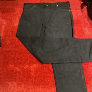 Svarta jeans från Wrangler 38/34 - Svarta jeans från Wrangler i klassisk rak modell. Byxorna har fem fickor, bälteshällor och detaljer i mässing. Tillverkade i 100% bomull för en skön och slitstark känsla. Snyggt läderpatch med Wrangler-logga baktill.