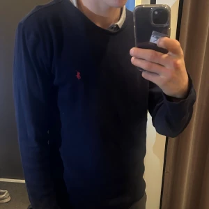 Mörkblå långärmad tröja Polo Ralph Lauren - Mörkblå långärmad tröja från Polo Ralph Lauren med klassisk röd broderad logga på bröstet. Tröjan är i ett ribbat bomullstyg och har en rund halsringning. Perfekt för dig som gillar stilren och enkel design.