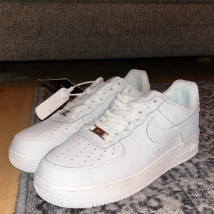 Nike Air Force 1 vita sneakers - Fräscha Nike Air Force 1 sneakers i helvitt med klassisk låg siluett. Skorna har perforerad tå, snörning och en platt sula med tydlig Air-dämpning. Tillverkade i skinn med vita detaljer och metallplatta på snörningen. Perfekta för dig som gillar clean och stilren look.