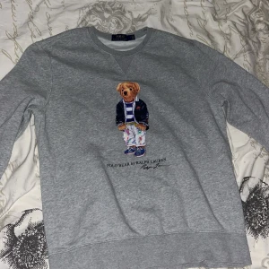 Grå sweatshirt Polo Bear Ralph Lauren - Grå sweatshirt från Polo Ralph Lauren med ikoniska Polo Bear-trycket framtill. Tröjan har rund halsringning och långa ärmar. Tillverkad i mjuk bomullsmix som är perfekt för chill dagar. Klassisk och stilren design med en touch av street.