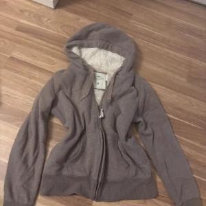 Brun hoodie med fuskpälsfoder H&M L.O.G.G. - Mysig brun hoodie från H&M L.O.G.G. med dragkedja och huva. Insidan är fodrad med fluffigt fuskpälsmaterial som gör den extra varm. Tröjan har långa ärmar, ribbade muddar och två fickor framtill. Perfekt för kyliga dagar.