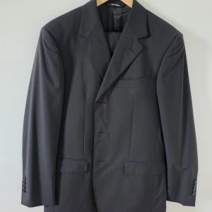 Mörkgrå randig kostym Ted Lapidus 46R - Snygg mörkgrå kostym från Ted Lapidus i storlek 46R. Kavajen har diskreta ränder, klassisk krage och knappar framtill. Byxorna matchar med samma mönster och har raka ben. Kostymen är tillverkad i ullblandning och passar dig som gillar stilren och tidlös look.