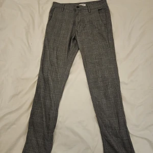 Grå rutig kostymbyxa Selected Homme - Snygga grå kostymbyxor från Selected Homme med diskret rutigt mönster. Byxorna har normal passform, bälteshällor och klassiska fickor fram och bak. Perfekt för dig som gillar stilrena och moderna detaljer.