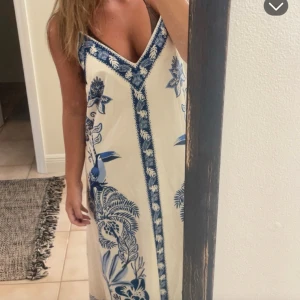 Vit och blå långklänning med v-ringning - Säljer en vit långklänning med blå tropiskt inspirerat blommönster och fågelmotiv. Klänningen har v-ringad hals och är ärmlös med smala axelband. Perfekt för varma dagar och har en luftig passform.
