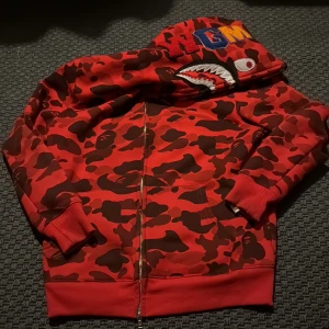 BAPE röd camo hoodie med hajmotiv - Säljer en ikonisk röd camo hoodie från BAPE med hajmotiv på huvan och broderade bokstäver. Tröjan har dragkedja hela vägen, känguruficka och ribbade muddar. Perfekt statement-plagg för dig som gillar streetwear.