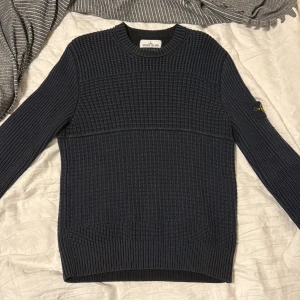 Mörkblå stickad tröja Stone Island - Säljer en mörkblå stickad tröja från Stone Island med klassisk patch på ärmen. Tröjan har grovstickad struktur, rund hals och ribbade muddar. Perfekt för dig som gillar streetwear och vill ha något både snyggt och bekvämt.