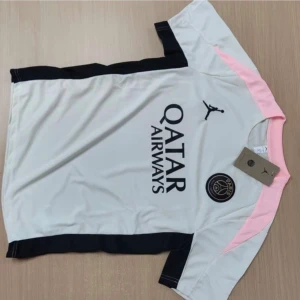 PSG x Jordan vit och svart fotbollströja - Snygg PSG x Jordan fotbollströja i vit med svarta och ljusrosa detaljer. Kortärmad modell med rund hals och tryckta loggor från Jordan och PSG på bröstet. Materialet är lätt och andas, perfekt för träning eller match. Säljes med matchande svarta shorts med rosa inslag och loggor.
