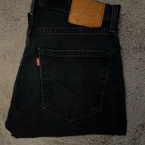 Levi's 502 mörkblå jeans W30 L32 - Mörkblåa Levi's 502 jeans med klassisk femficksdesign och taper fit. Jeansen har mörk tvätt, diskreta slitningar och den ikoniska läderpatchen bak i midjan. Perfekta för dig som gillar en clean och tidlös look. Pyttelitet hål på ena knäet.