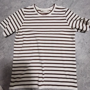 Randig t-shirt från Wear Copenhagen XS - Snygg t-shirt från Wear Copenhagen 1969 i storlek XS. T-shirten är vit med bruna horisontella ränder och har klassisk rund hals. Tillverkad i mjuk bomull och har korta ärmar. Perfekt för en chill och stilren look.