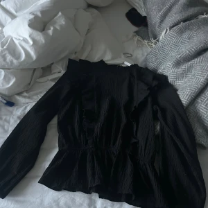 Svart blus med volangdetaljer - Säljer en svart blus med långärmade och volangdetaljer längs axlar och bröst. Blusen har en lätt skrynklig struktur och rund hals med liten knapp i nacken. Passformen är normal och materialet känns som polyester. Perfekt för dig som gillar stilrena och trendiga plagg.
