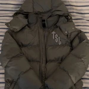 Grå pufferjacka Trapstar London S - Säljer en grå pufferjacka från Trapstar London med stor broderad logga och pantermotiv på ryggen samt TSL-tryck på bröstet. Jackan har huva, dragkedja och är riktigt varm och street. Perfekt för kalla dagar. Storleken är S på jackan men passar även M