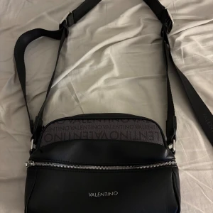 Svart axelväska från Valentino - Snygg svart axelväska från Valentino med justerbar rem och silverfärgad dragkedja. Väskan har logotypmönster upptill och är tillverkad i skinnliknande material. Perfekt storlek för det viktigaste och stilren design med diskret branding framtill.