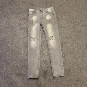 Grå slitna jeans med skinny fit - Snygga grå jeans med slitningar och trasiga detaljer på framsidan. Skinny fit och klassisk femficksmodell. Materialet är 85% bomull och 15% polyester, vilket gör dem både mjuka och hållbara. Perfekt för dig som gillar en cool och avslappnad stil.