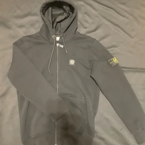 Svart hoodie från Stone Island - Svart hoodie från Stone Island med dragkedja, huva med snörning och klassisk logotyp på bröstet samt ikonisk patch på ärmen. Tröjan har känguruficka och är tillverkad i mjuk bomull. Perfekt för dig som gillar streetwear och vill ha en clean look.