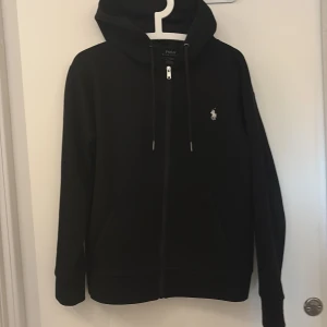Svart hoodie från Polo Ralph Lauren - Svart hoodie från Polo Ralph Lauren med klassisk logga broderad på bröstet. Tröjan har huva med snörning, dragkedja framtill och två fickor. Långärmad och tillverkad i mjuk bomull, perfekt för chill dagar.