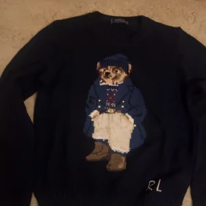 Mörkblå stickad tröja Polo Bear - Säljer en mörkblå stickad tröja från Polo Ralph Lauren med ikoniska Polo Bear-motivet framtill. Tröjan har rund hals, långa ärmar och RL-broderi vid nederkanten. Perfekt för dig som gillar klassisk preppy stil med en twist.