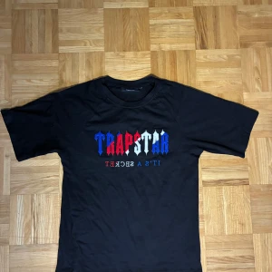 Trapstar svart t-shirt med brodyr - Svart t-shirt från Trapstar med broderad logga i blått, rött och vitt på bröstet. Klassisk passform med rund hals och korta ärmar. Materialet är mjuk bomull och trycket har en fet streetkänsla. Perfekt för dig som vill sticka ut med en ikonisk look.