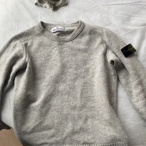 Grå stickad tröja från Stone Island - Snygg grå stickad tröja från Stone Island Junior med klassisk rund hals och ikonisk logga på ärmen. Tröjan är tillverkad i en mjuk blandning av polyamid och 10% kashmir för extra komfort. Perfekt för dig som gillar stilrena och exklusiva plagg.