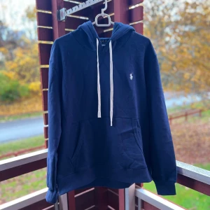 Mörkblå hoodie från Polo Ralph Lauren - Snygg mörkblå hoodie från Polo Ralph Lauren med vit snörning och klassisk logga broderad på bröstet. Tröjan har huva, känguruficka och ribbade muddar. Perfekt för chill dagar och enkel att matcha med jeans eller joggers.