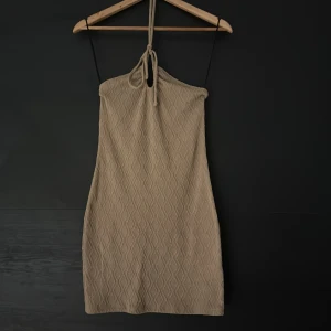 Beige stickad kortklänning med snörning - Supersnygg beige kortklänning med mönster och knyt i nacken som kan även användas som en bandeau klänning. Klänningen är ärmlös och har en slim passform som framhäver siluetten. Perfekt för dig som gillar en enkel men trendig look med lite textur och detaljer. 