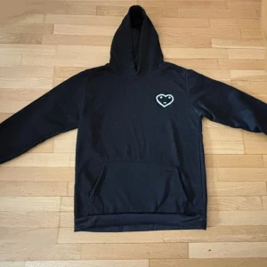 Carsiko set - Svart hoodie med huva och magficka. På bröstet finns en broderad hjärtdetalj med ett litet ansikte i vitt. Hoodien har långa ärmar och en klassisk, avslappnad passform. Perfekt för dig som gillar stilrena plagg med en söt twist.