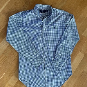 Rutig skjorta från Ralph Lauren S - Snygg rutig skjorta från Ralph Lauren i klassisk passform. Skjortan har blå, gröna och vita rutor, långärmad design och knappar framtill. Tillverkad i mjuk bomull och har den ikoniska Polo-loggan broderad på bröstet. Perfekt för dig som gillar stilrena och tidlösa plagg.