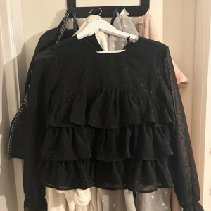 Svart volangblus från Gina Tricot - Supersnygg svart blus från Gina Tricot med flera lager volanger framtill och diskreta prickar i tyget. Blusen har långärmade transparenta ärmar och en loose passform. Perfekt för dig som gillar detaljer och vill sticka ut lite extra.