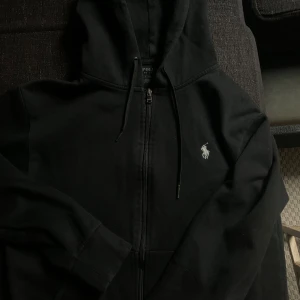 Svart hoodie från Polo Ralph Lauren - Svart hoodie från Polo Ralph Lauren med dragkedja framtill och klassisk huva med snörning. Tröjan har lång ärm och den ikoniska broderade loggan i vitt på bröstet. Tillverkad i mjuk bomull, perfekt för chill dagar.