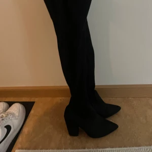 Mocka höga stövlar med klack - Säljer ett par svarta mocka höga stövlar med chunky klack och spetsig tå. Skorna är i mockaliknande material och går upp över knäet för en riktigt snygg siluett. Perfekta för att lyfta vilken outfit som helst.