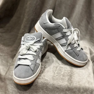Grå Adidas Campus sneakers i mocka - Snygga Adidas Campus sneakers i grå mocka med vita detaljer och klassiska tre ränder på sidorna. Skorna har vit sula med gummiyta undertill och breda vita skosnören. Perfekt för dig som gillar retrovibbar och streetstyle.