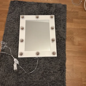 Sminkspegel med LED-belysning - Elegant sminkspegel med 10 LED-lampor runt ramen för optimal belysning. Spegeln är vit och ansluts med strömkabel, perfekt för makeup eller styling. Lamporna fungerar och ger starkt ljus. Mindre slitage syns på ramen, men i gott skick.