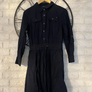 Marinblå skjortklänning Tommy Hilfiger - Snygg marinblå skjortklänning från Tommy Hilfiger med knappar hela vägen framtill, krage och två bröstfickor. Klänningen har lång ärm, knytband i midjan och är tillverkad i mjuk bomull. Perfekt för dig som gillar klassisk och stilren design.